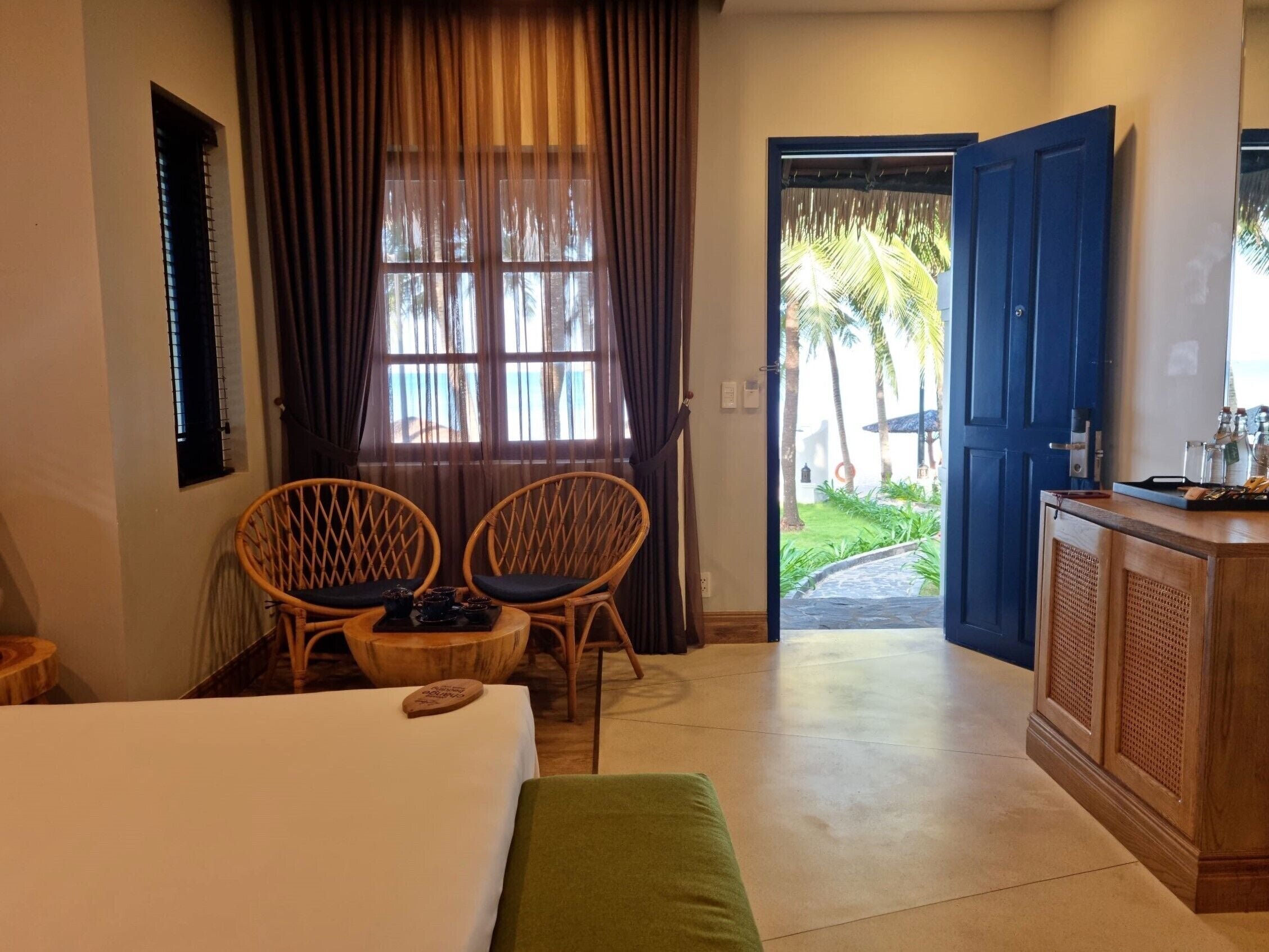 Quarto de hotel com cama, duas cadeiras de vime, uma mesinha com bandeja, um armário de madeira e vista para um jardim através de uma porta aberta.