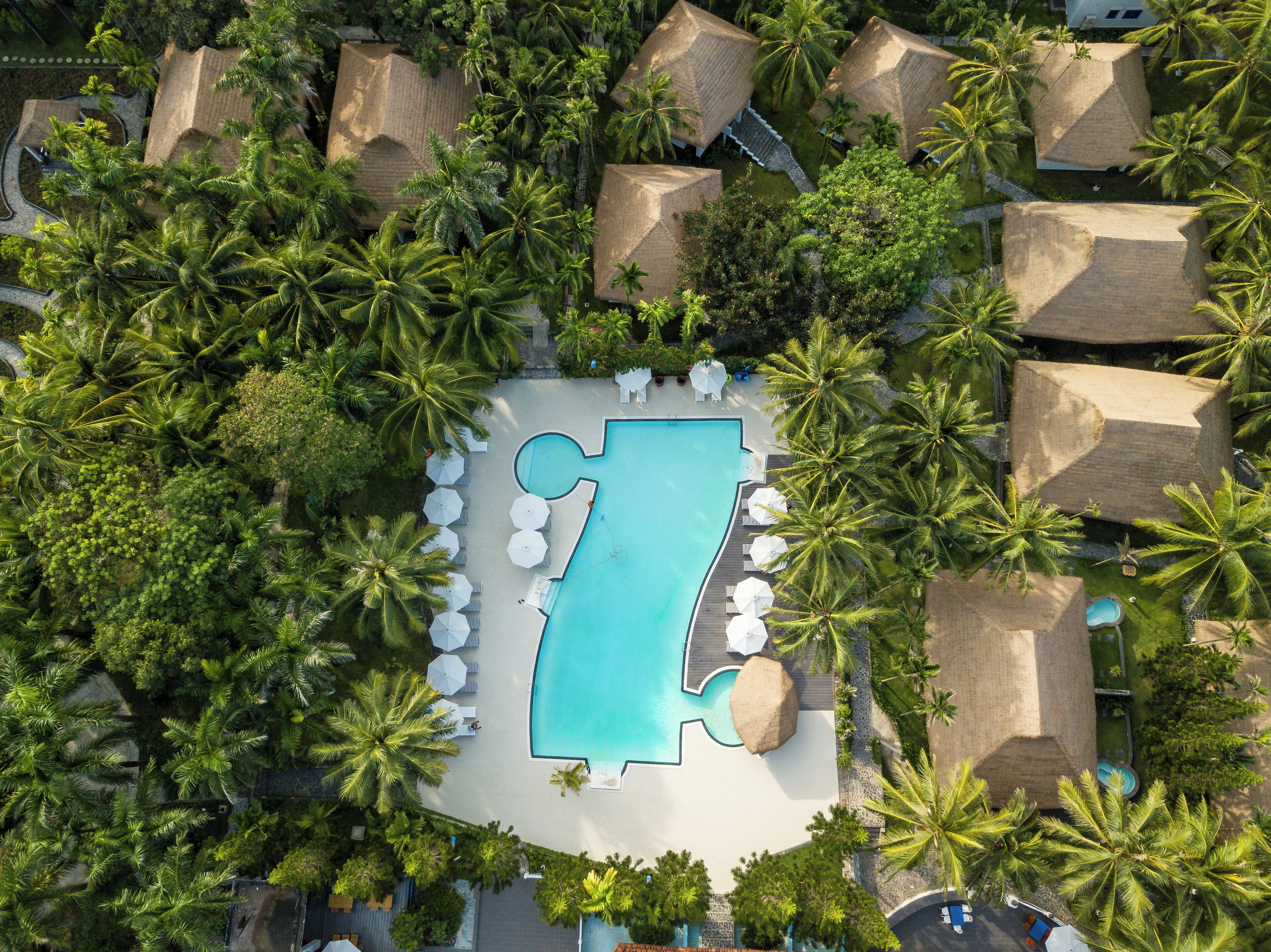An aerial view of a resort with a large pool, palm trees, and thatched-roof huts.