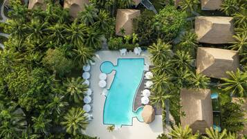 An aerial view of a resort with a large pool, palm trees, and thatched-roof huts.
