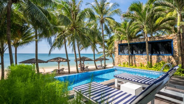 A pool area with lounge chairs, a stone wall, and a view of the beach and ocean. - L'Azure Resort and Spa (Phu Quoc)