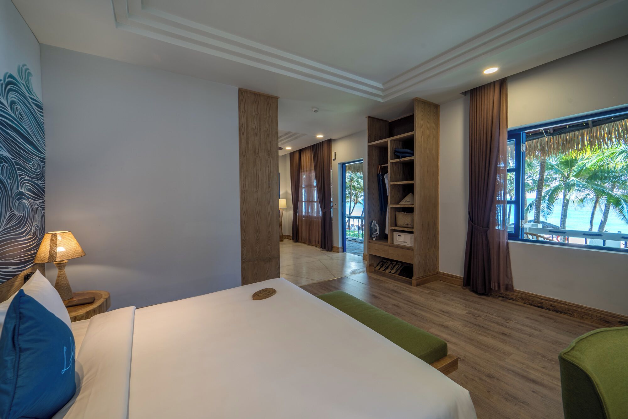 A hotel room with a large bed, a wardrobe, a vanity with a mirror, a window with a view of palm trees, and a door leading to another room.