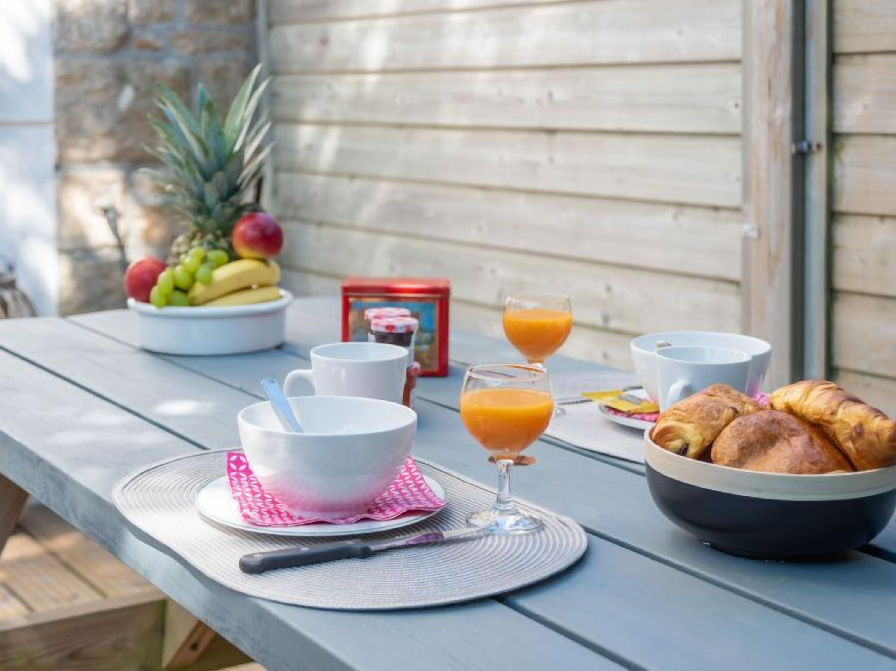 A breakfast setup on an outdoor table with a bowl of fruit, two glasses of orange juice, and a bowl of croissants.