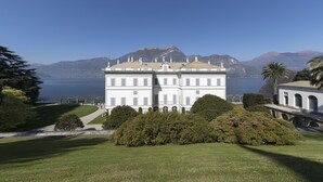 A large, white building with multiple windows and a central tower, surrounded by lush greenery and mountains in the background. - Villa Mojana, Limonta, Bellagio | The D’Angelo Family Collection (Oliveto Lario)