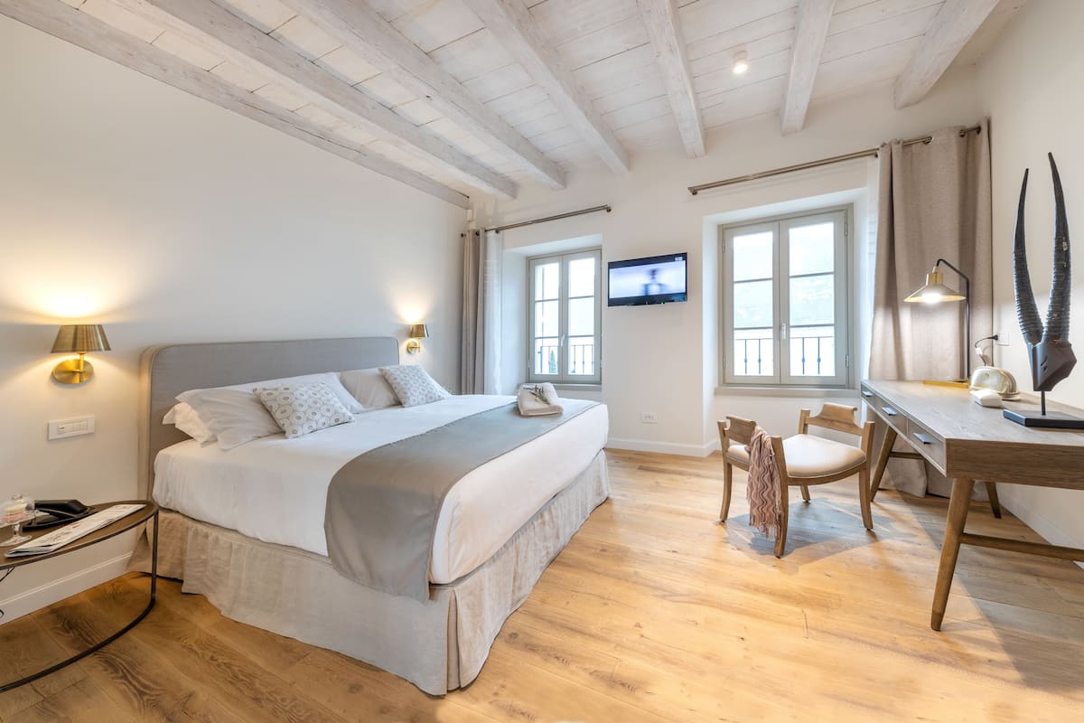 A bedroom with a bed, bedside table, lamp, TV, desk, chair, and wooden floor.
