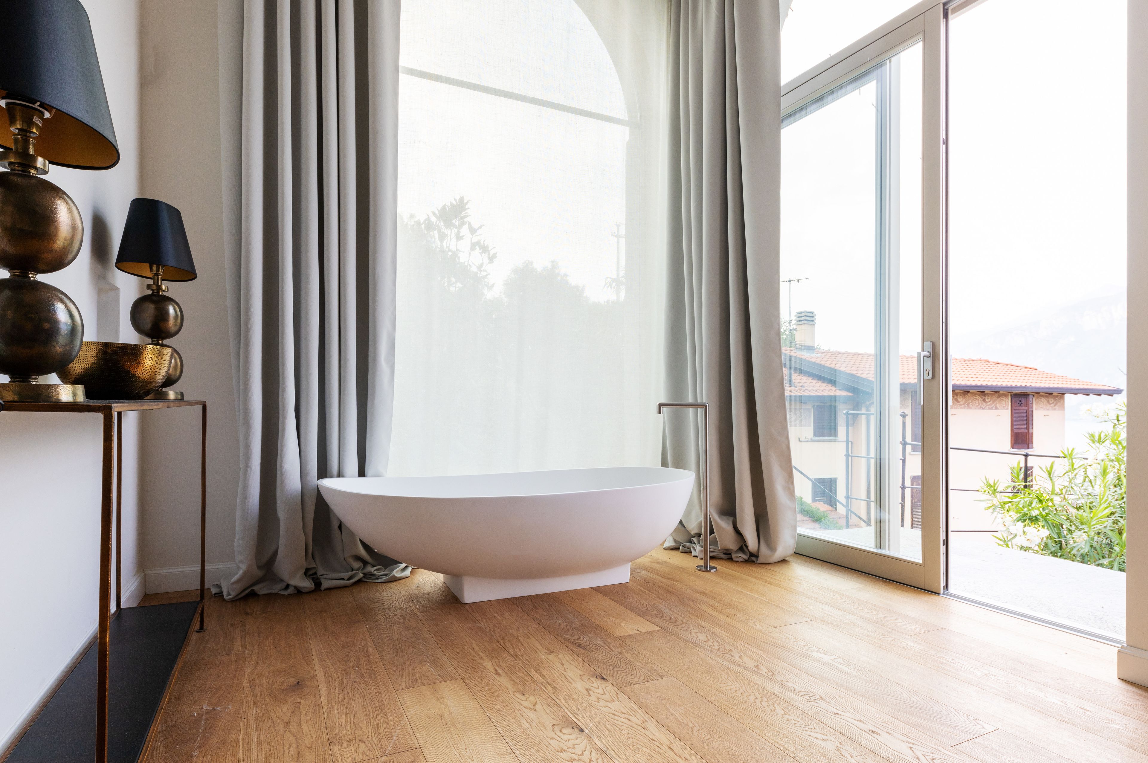 Un bagno moderno con una vasca freestanding, pavimento in legno e un'ampia finestra con tende.