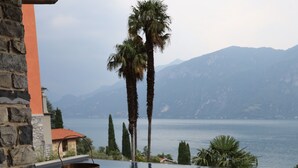 A wooden table with a candle, a cushioned chair, and a view of a swimming pool with palm trees and mountains in the background. - Villa Mojana, Limonta, Bellagio | The D’Angelo Family Collection (Oliveto Lario)
