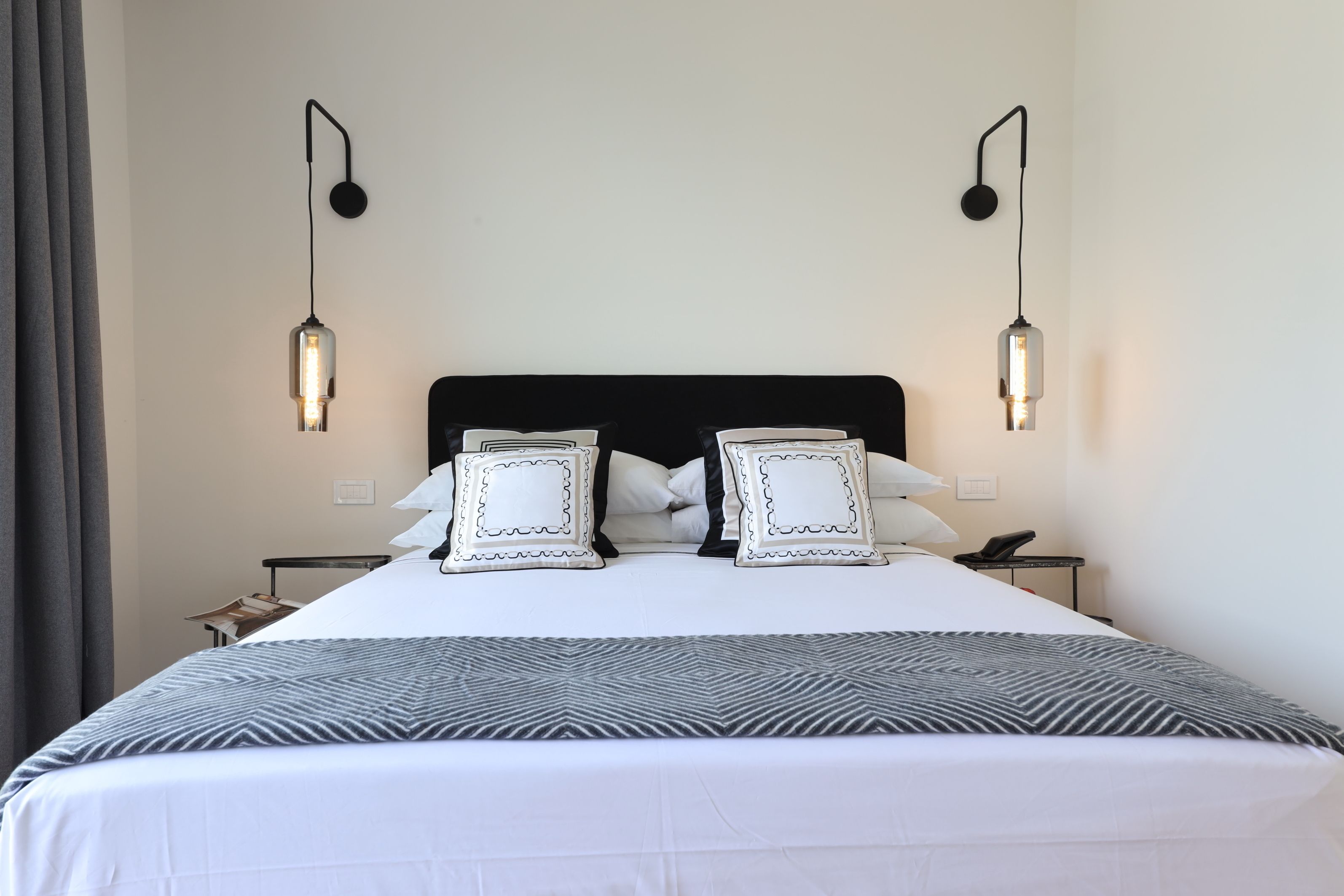 A neatly made bed with a black headboard, white bedding, and decorative pillows. There are two bedside tables with lamps and a small table with a telephone.