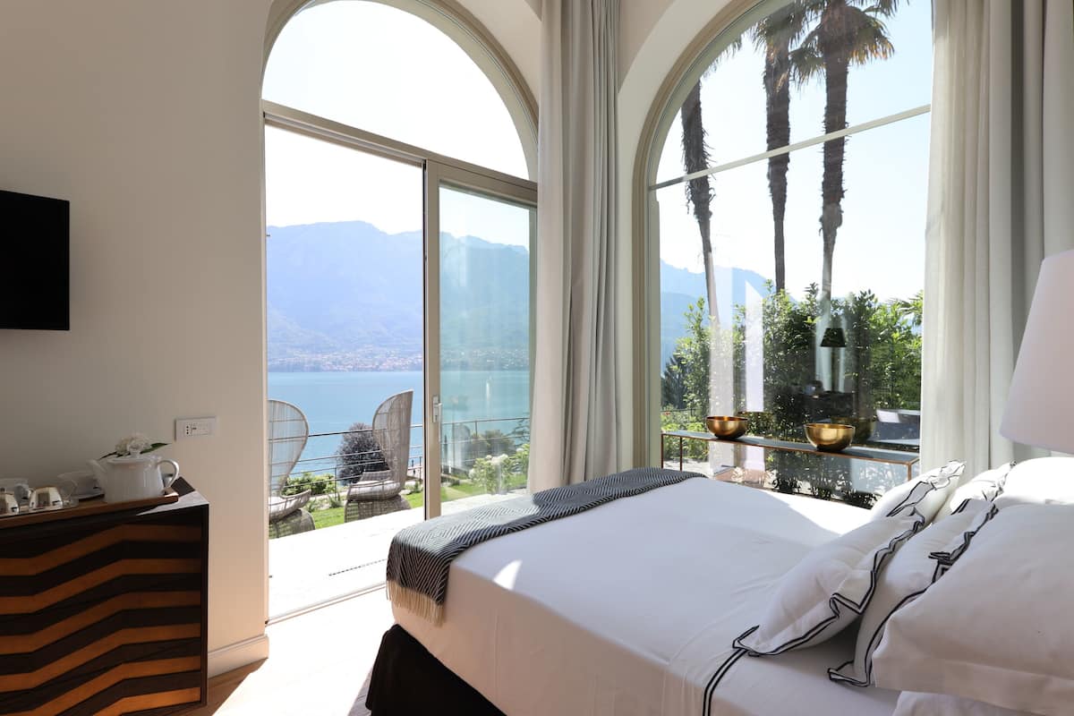 A bedroom with a large bed, a TV mounted on the wall, and a balcony with a view of the sea and mountains.