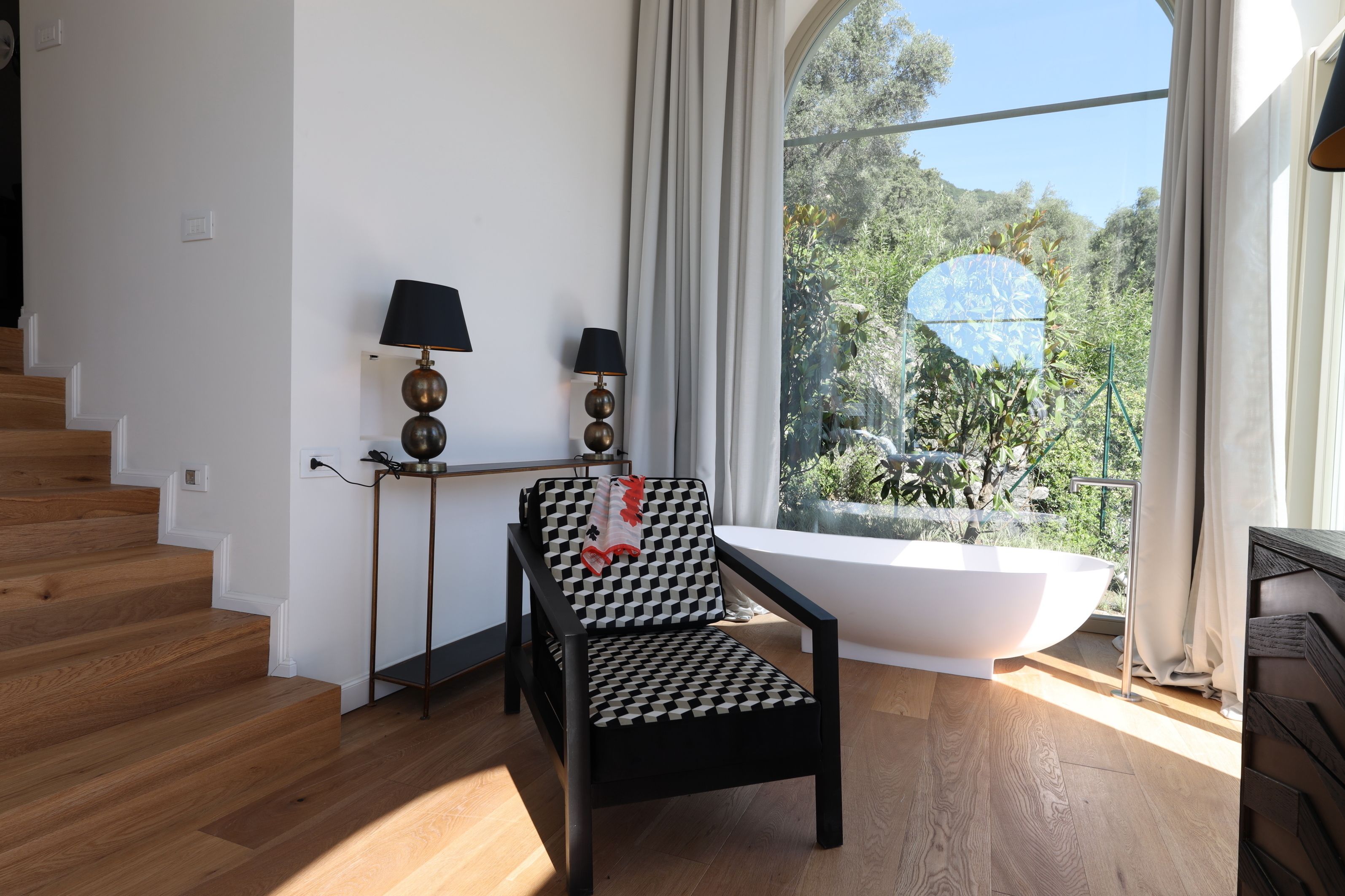 Un bagno moderno con una vasca freestanding, una sedia a scacchi bianchi e neri, e un'ampia finestra con vista su del verde.