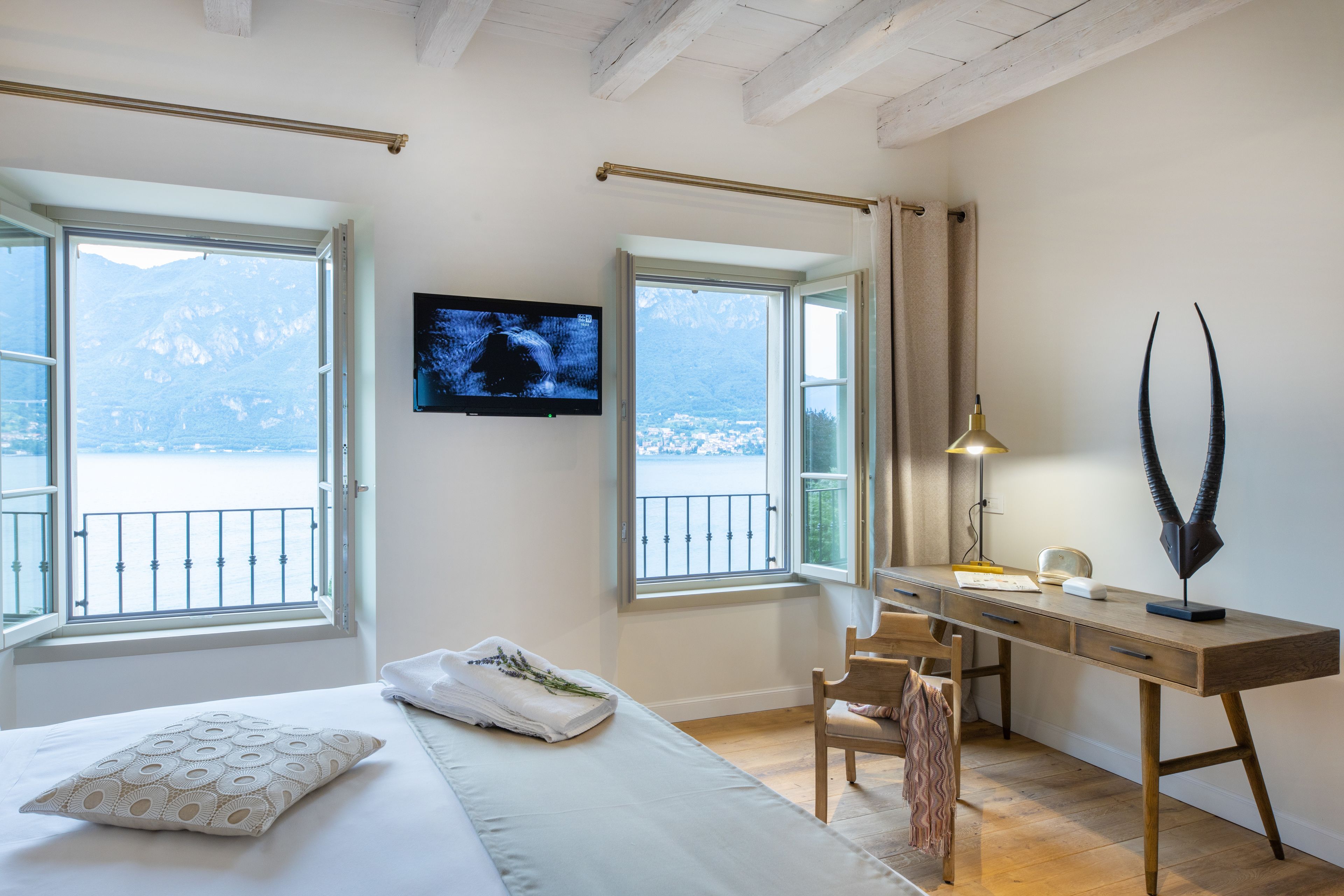 A bedroom with a bed, a desk, and a TV. There are windows with a view of mountains and a balcony.