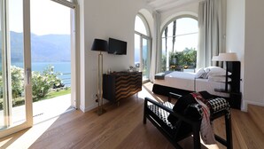 A bedroom with a bed, a desk, a chair, a dresser, and a TV. There is a large window with a view of the mountains and the sea. - Villa Mojana, Limonta, Bellagio | The D’Angelo Family Collection (Oliveto Lario)