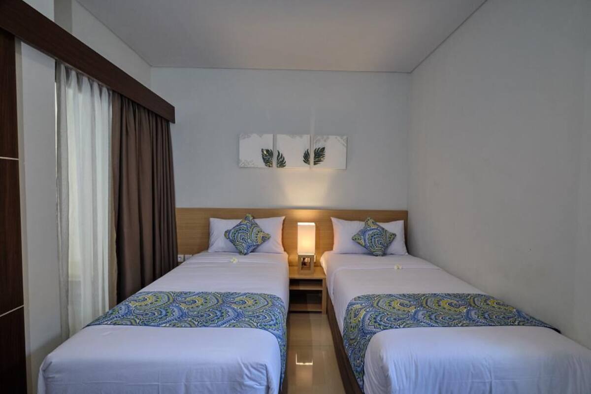 Ein Hotelzimmer mit zwei Einzelbetten, einem Nachttisch mit Lampe und einem Fenster mit Vorhängen.