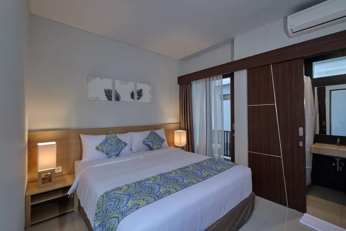 Ein Hotelzimmer mit einem großen Bett, einem Nachttisch mit Lampe, einem Badezimmer mit Waschbecken und einem Fenster mit Vorhängen.