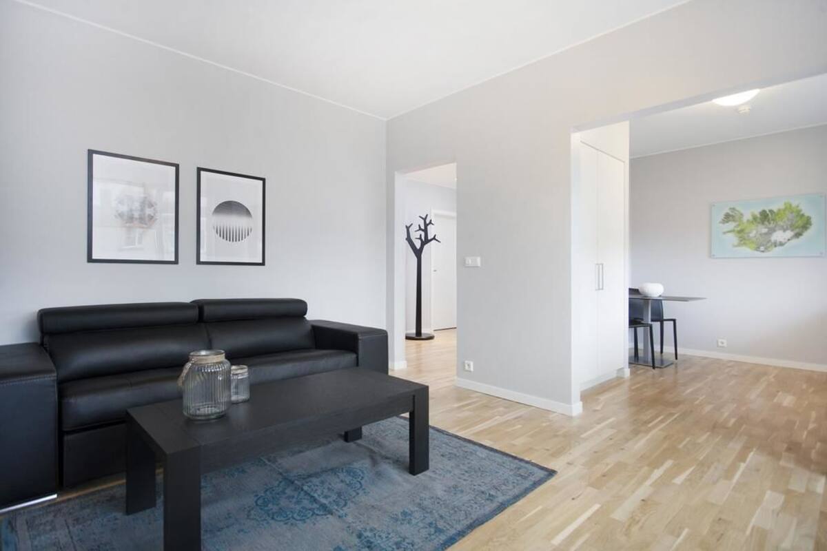 Un salon moderne avec un canapé en cuir noir, une table basse sombre et un tapis à motifs bleus. Deux œuvres d’art encadrées ornent le mur, et une peinture est accrochée au mur adjacent.