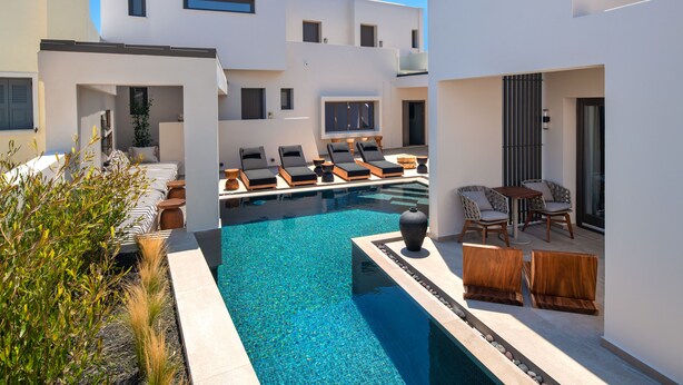 Un área de piscina moderna con sillones de descanso y una pequeña mesa, rodeada de edificios blancos con balcones.