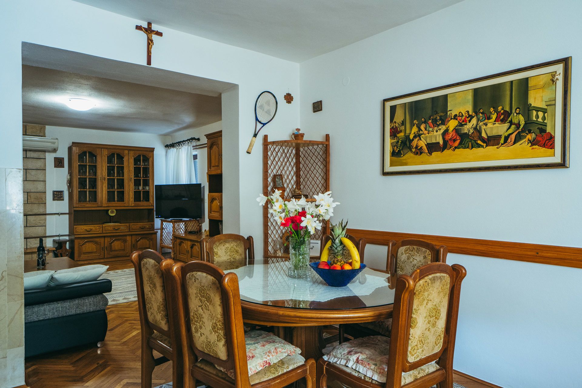 A dining area with a round table, chairs, a painting, and a cross on the wall.