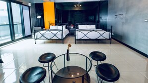 Una habitación de hotel moderna con dos camas, una mesa de vidrio y cuatro taburetes, y un ventanal que ofrece vistas al exterior.