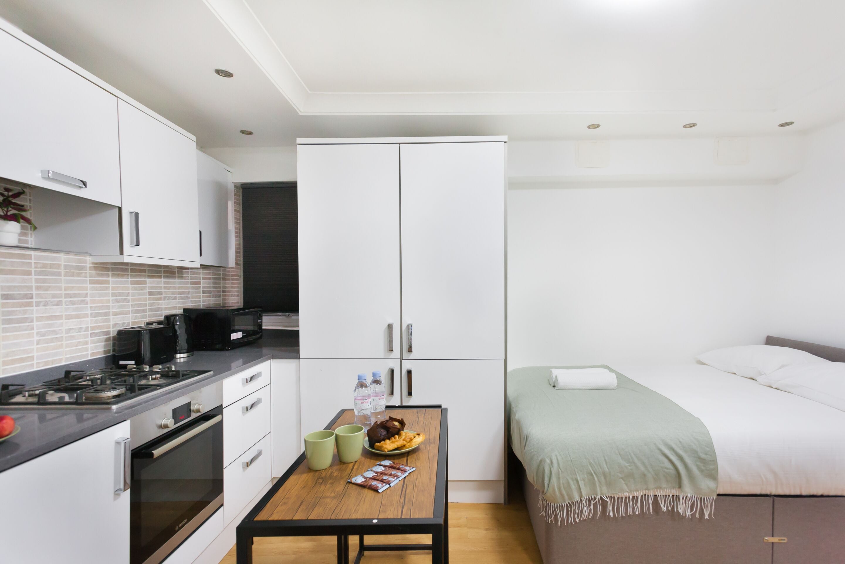 A compact kitchen with a stove, oven, and microwave, a dining table with a plate of food and drinks, and a bed with a green blanket.