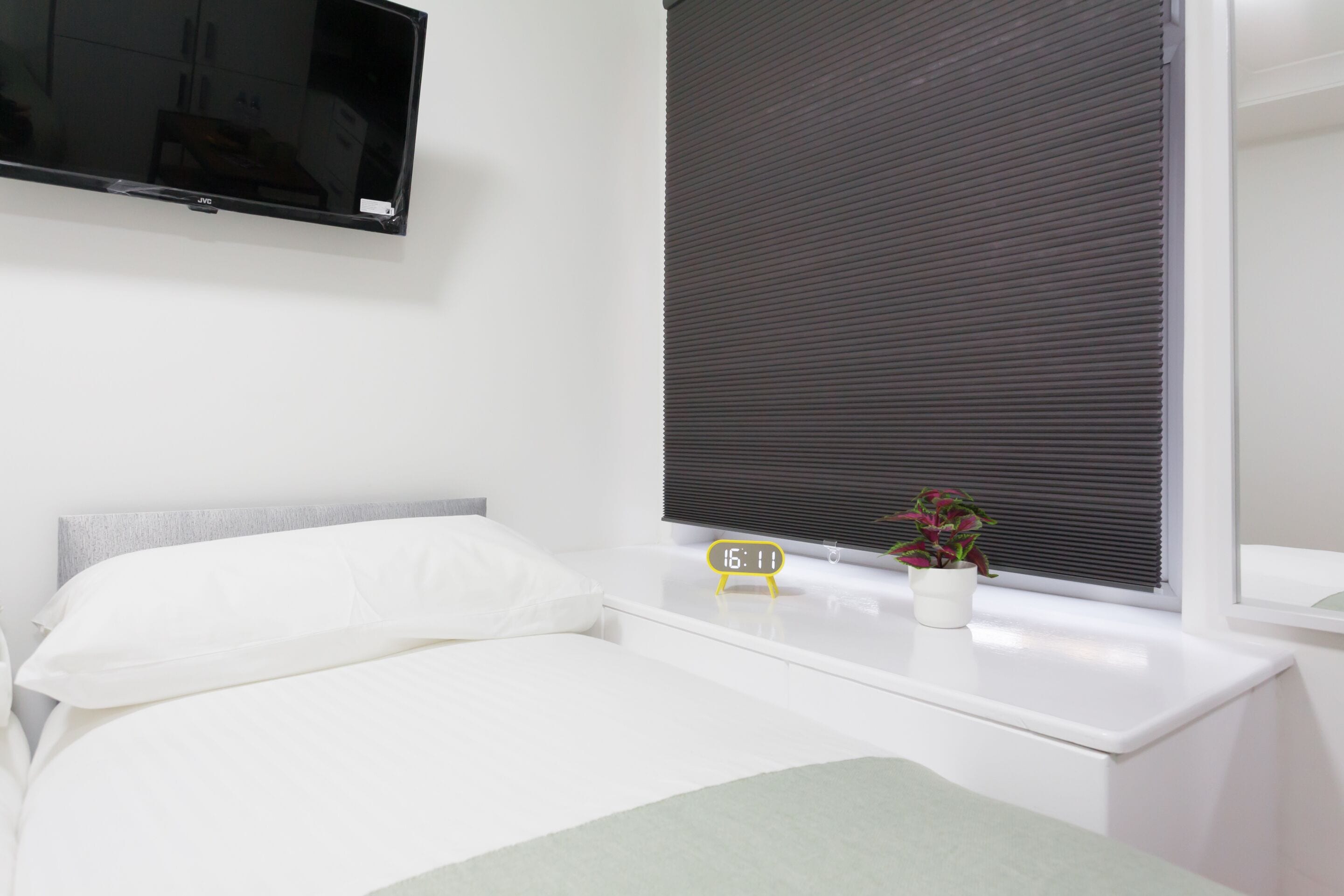 A modern bedroom with a bed, a bedside table with a clock, a potted plant, and a window with blinds.