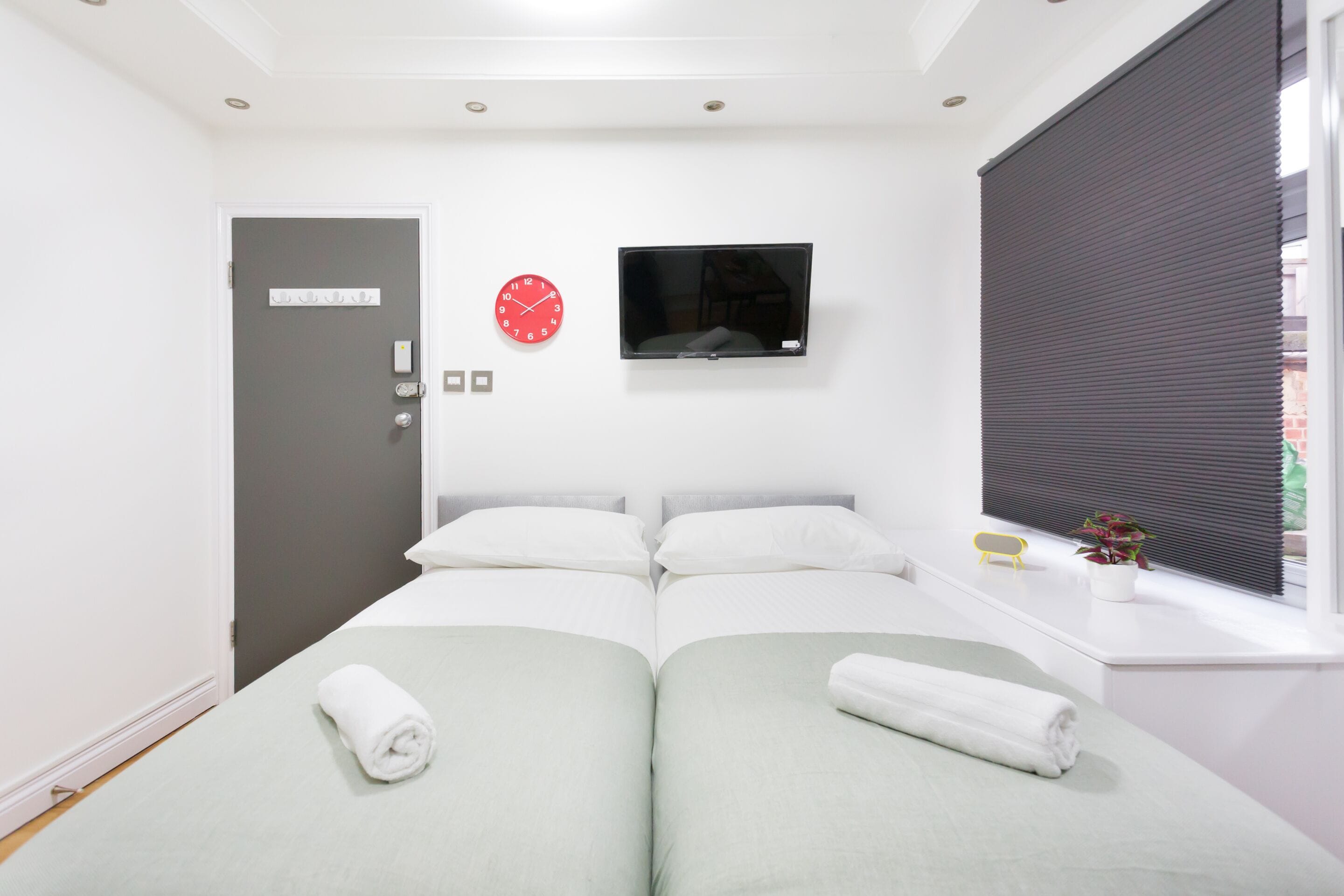 A hotel room with two beds, a TV mounted on the wall, a red clock, a door with a control panel, and a window with blinds.