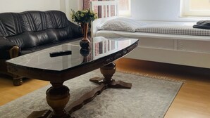 A living room with a black leather sofa, a glass-top coffee table with a remote control, a white cushioned bench, and a window with sheer curtains. - City Pension Bremerhaven (Bremerhaven)