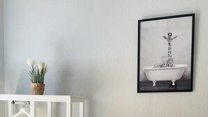 A room with a white shelving unit, a framed picture of a giraffe in a bathtub, and a basket with plants on the wall. - City Pension Bremerhaven (Bremerhaven)
