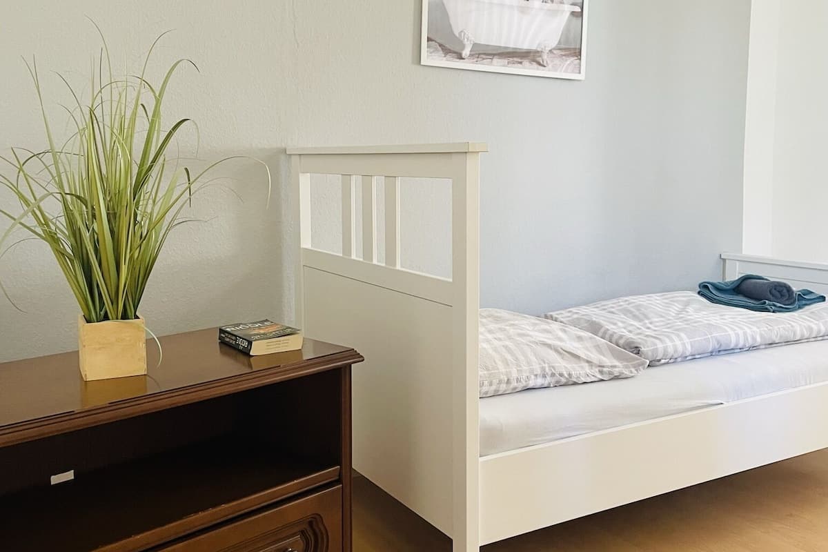 A bedroom with a white bed, a wooden dresser, a potted plant, and a framed picture on the wall.