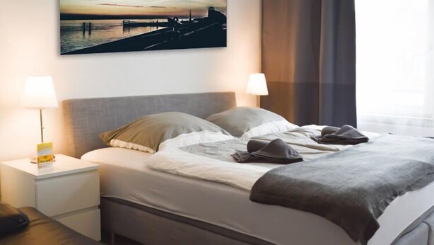A neatly made bed with pillows and a blanket, a bedside table with a lamp, and a painting on the wall. - City Pension Bremerhaven (Bremerhaven)