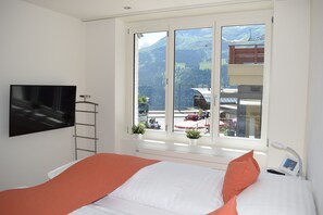 A bedroom with a bed, a TV, a window with a mountain view, and a small table with a lamp. - Apartement Eigerpeak (Lauterbrunnen)