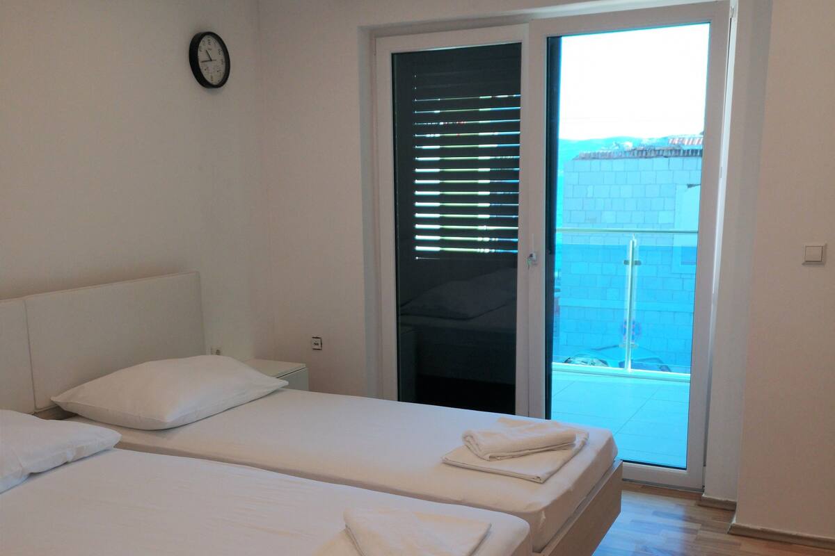 A bedroom with two beds, a clock on the wall, and a sliding glass door leading to a balcony.