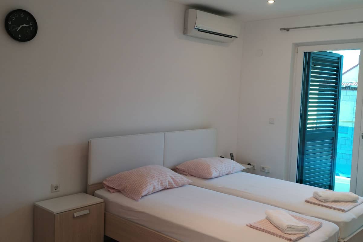 A bedroom with two beds, a wall clock, an air conditioning unit, and a window with blinds.