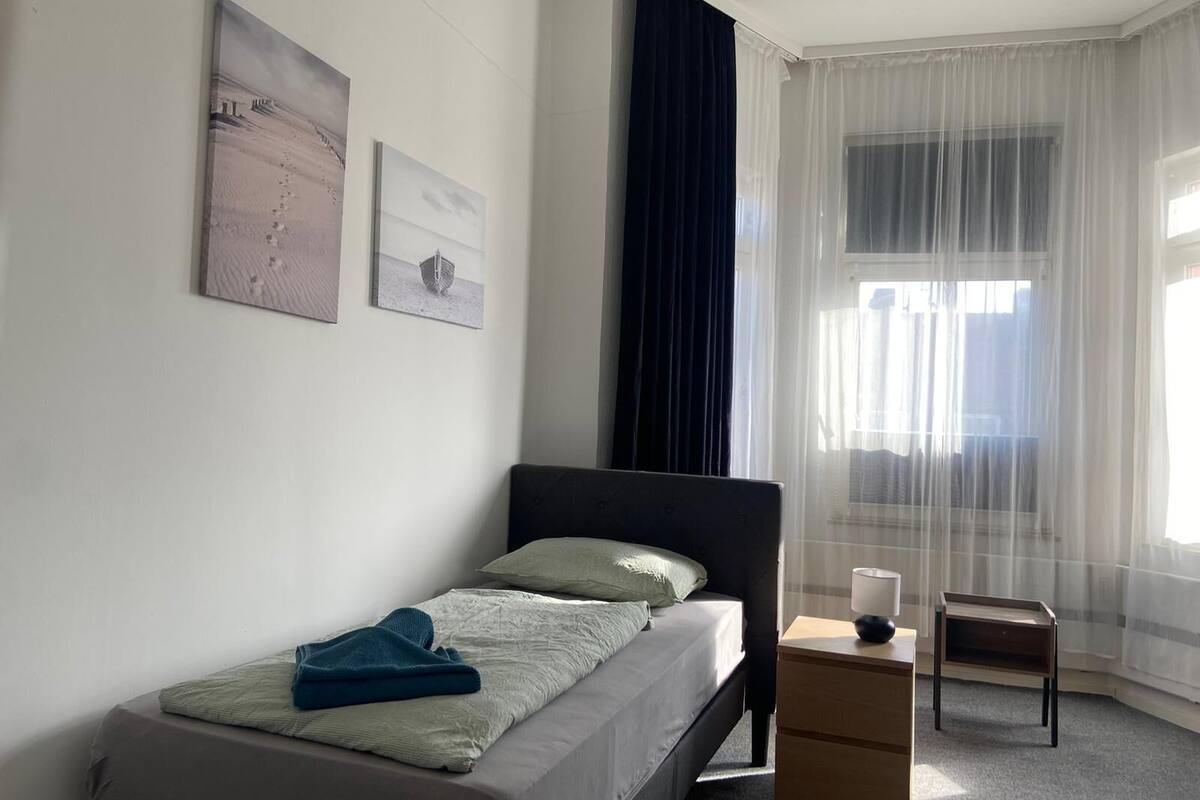 Une chambre à coucher avec un lit, une table de chevet, une lampe et une fenêtre avec des rideaux légers.