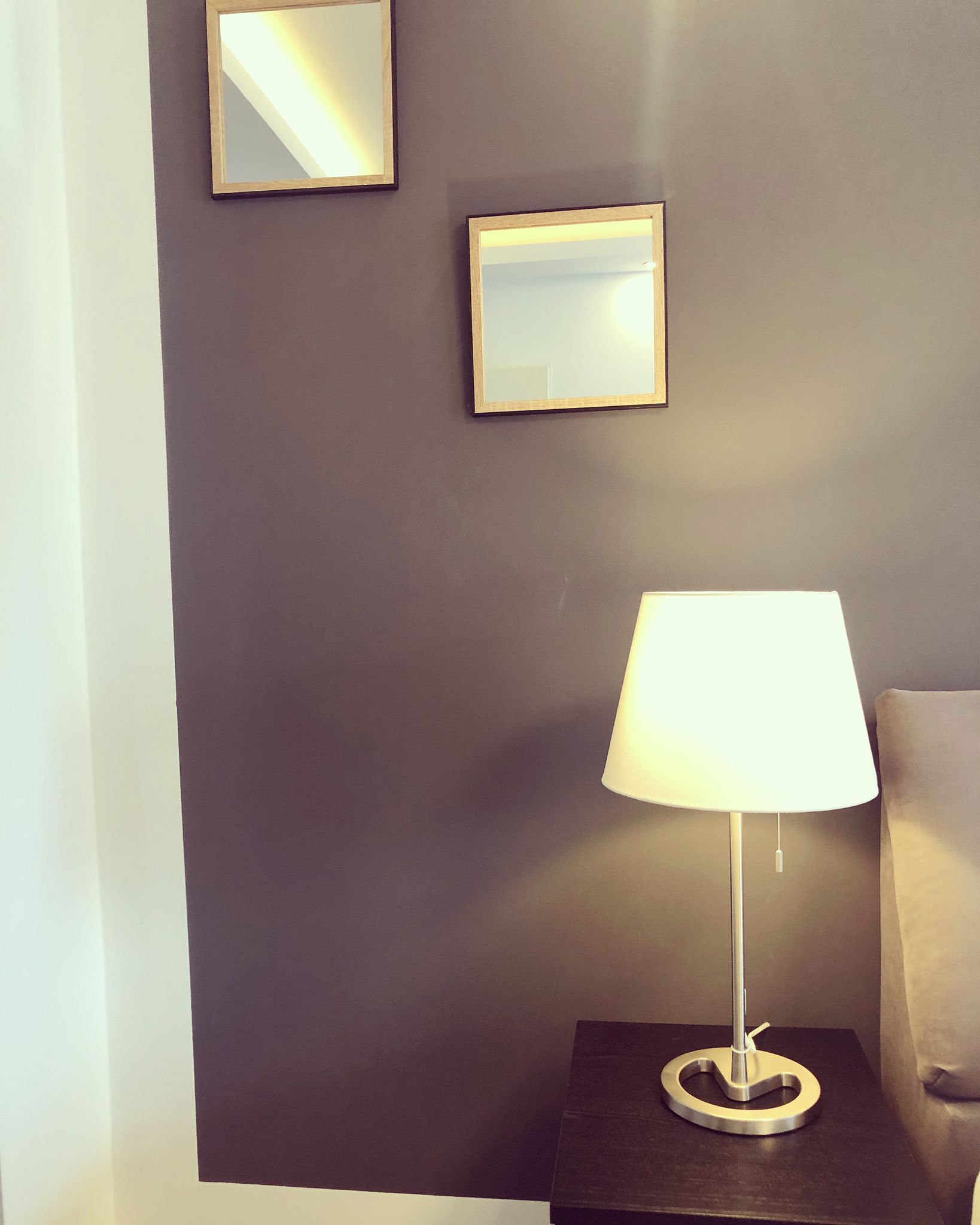 A room with a dark wall, a small table with a lamp, and two framed mirrors.
