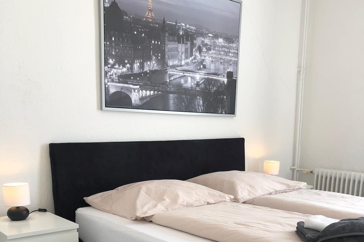 Une chambre à coucher avec une tête de lit noire, du linge de lit blanc, une table de chevet avec une lampe et un tableau représentant un paysage urbain encadré accroché au mur.