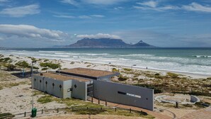 Un edificio costiero con tetto piatto, situato vicino a una spiaggia con onde e una montagna in lontananza.