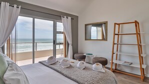 Una camera d'albergo con un letto grande, vista sull'oceano, una scaffalatura a scaletta in legno e un tavolino rotondo.