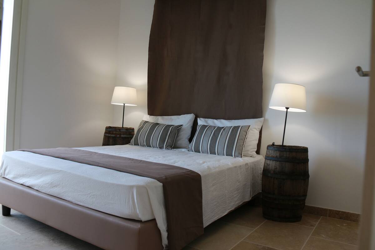 Una cama bien hecha con un cabecero marrón, dos lámparas de noche y un barril de madera al lado de la cama.
