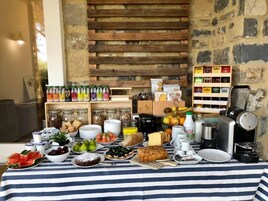 A breakfast buffet with various food items and beverages, including fruits, pastries, and condiments, set against a rustic stone wall backdrop.