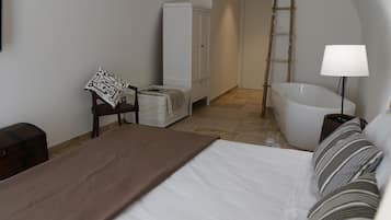 Un dormitorio con cama, una silla, armario, bañera y lámpara de noche.