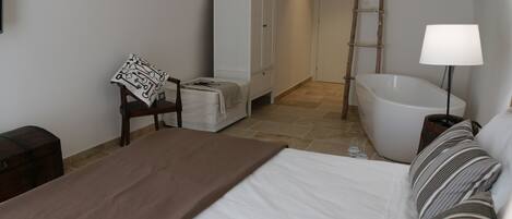 A bedroom with a bed, a chair, a wardrobe, a bathtub, and a bedside lamp.