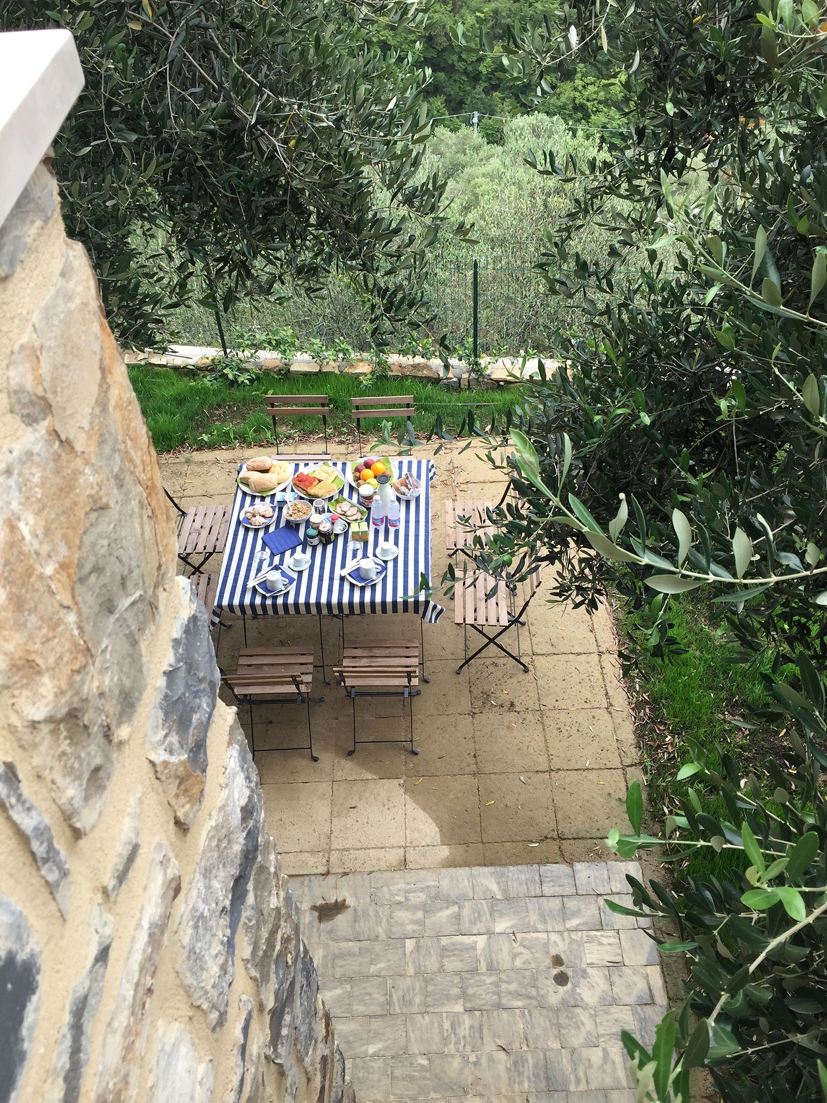 An outdoor dining area with a table set for two, surrounded by greenery and a stone wall.