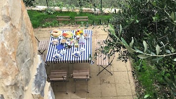 An outdoor dining area with a table set for two, surrounded by greenery and a stone wall.