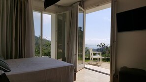 Un dormitorio con cama, una puerta corrediza de vidrio que da a un balcón con una silla y vistas a vegetación y una cerca.