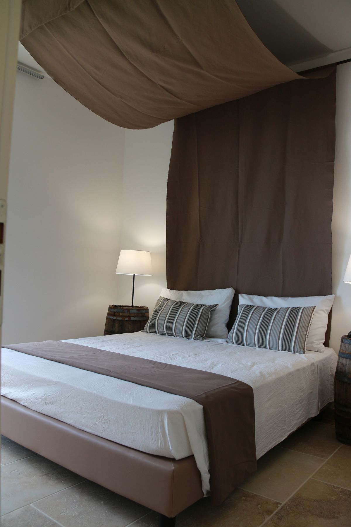 Un dormitorio con una cama grande, dosel, mesita de noche con lámpara y un barril de madera.