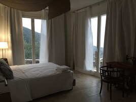 A hotel room with a bed, a table, and chairs. There are curtains and a view of the outdoors.