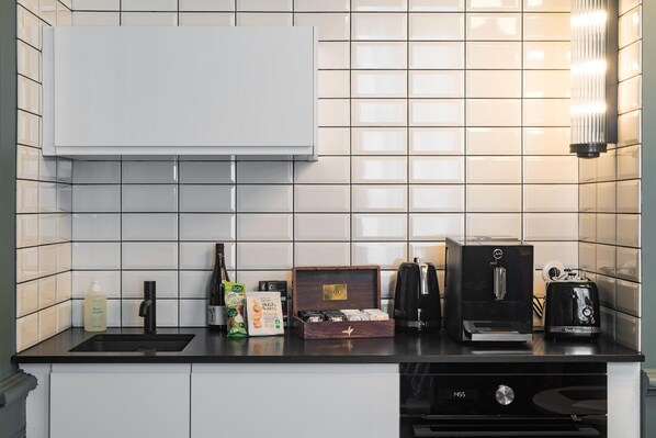 A modern kitchen with white tiles, a black countertop, and various appliances including a coffee maker, toaster, and a box of snacks. - Villa 216 (Lyon)