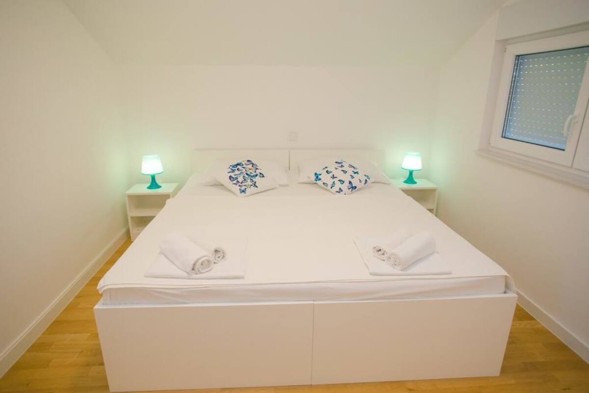A single-bed room with a white bed, two nightstands with lamps, and a window with blinds.