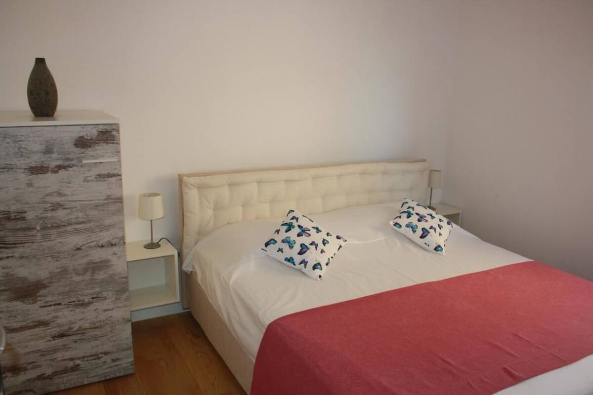 A bedroom with a white bed, two patterned pillows, a red blanket, a wooden dresser, and a bedside lamp.