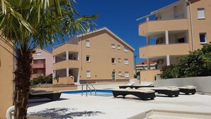 A pool area with lounge chairs, a palm tree, and multi-story buildings in the background. - Apartments Orange (Novalja)