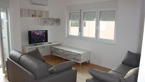 A modern living room with a flat-screen TV, grey sofas, a white console table, and a window with blinds. - Apartments Orange (Novalja)
